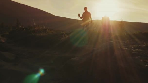 Silhouetten van een man en een vrouw met rugzakken en trekking stokken naar beneden het bergpad. In de stralen van de ondergaande zon — Stockvideo