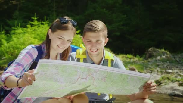 Ett ungt par turister studerar tillsammans en karta. De sitter på en naturskön plats nära en mountain river. 4k-video — Stockvideo