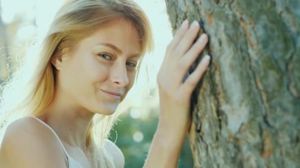 Portrait d'une belle jeune femme debout près de l'arbre au soleil, souriant, regardant la caméra. Communication avec la nature — Video
