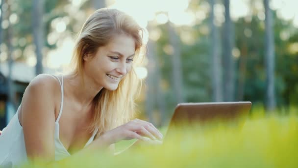 Een aantrekkelijke Kaukasische vrouw spreekt via de online chat. Ligt op het groene gras in de achtertuin van het huis — Stockvideo