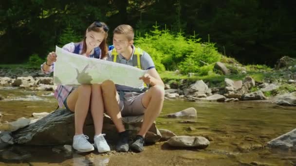 Una joven pareja de turistas estudian juntos un mapa. Se sientan en un lugar pintoresco cerca de un río de montaña. Composición del espacio de copia — Vídeos de Stock