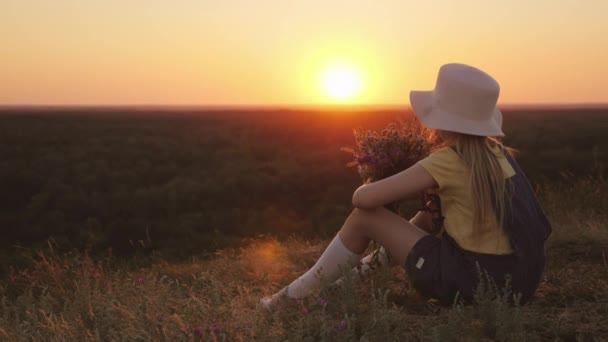 Το παιδί θαυμάζει το ηλιοβασίλεμα. Κάθεται πάνω σε ένα λόφο, κρατώντας ένα μπουκέτο αγριολούλουδα. Καλοκαίρι στο χωριό της έννοιας — Αρχείο Βίντεο