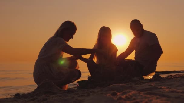 Los padres juegan con la hija en la arena cerca del mar, construyen juntos un castillo de arena — Vídeos de Stock