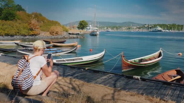 一个背着背包的妇女坐在挪威奥斯陆城市的海滨, 靠近渔船。使用智能手机。周游斯堪的纳维亚国家 — 图库视频影像