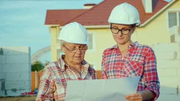 Uma mulher idosa com sua filha adulta inspecionando o novo prédio é verificada contra o plano de construção — Vídeo de Stock