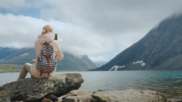 Een vrouw neemt een foto van een schilderachtig uitzicht op een hoge-bergachtige Noorse meer. Vakantie aan de rand van de wereld — Stockvideo
