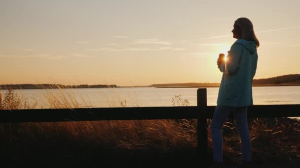 Una donna sola beve birra da una lattina sul lago. In piedi da solo vicino alla recinzione, guardando il tramonto — Video Stock