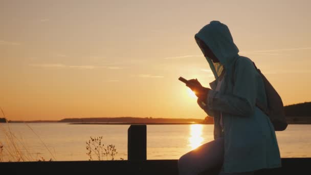フードの女性は絵のように美しい湖の近くのフェンスに座って、スマートフォンを使用しています。美しい夕日 — ストック動画