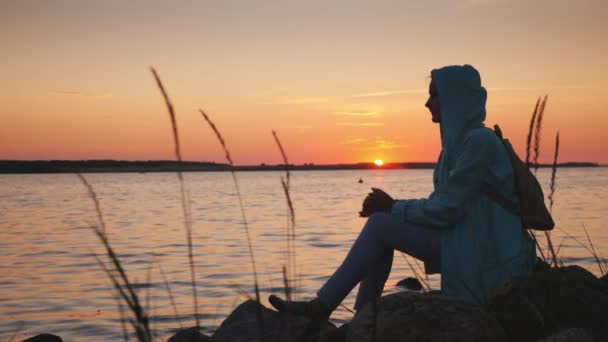 Μια μοναχική γυναίκα κάθεται σε έναν βράχο κοντά σε μια γραφική λίμνη στο ηλιοβασίλεμα — Αρχείο Βίντεο