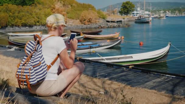 Achteraanzicht van een vrouw rust door de zee met uitzicht op de stad van Oslo, met behulp van een smartphone — Stockvideo