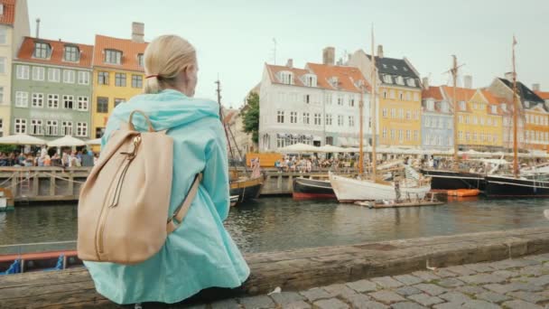Uma mulher se senta no aterro e admira os edifícios coloridos nas margens do canal Nyhavn em Copenhague — Vídeo de Stock