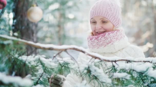 Жінка середнього віку в в'язаному теплому капелюсі прикрашає новорічну ялинку в засніженому лісі — стокове відео