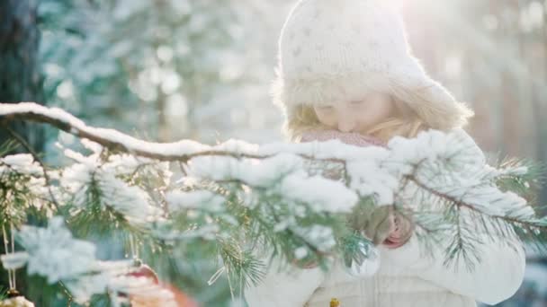 Het kleine meisje in een warme gebreide muts bereidt voor Kerstmis, siert de nieuwe jaar boom met kleurige ballen in de tuin van het huis. Op de takken witte sneeuw glitters — Stockvideo