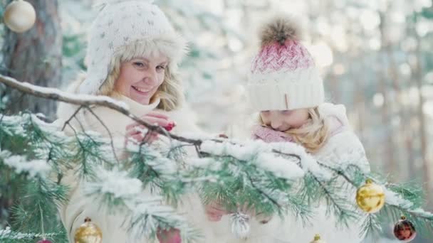 Een klein meisje en een jonge moeder decoreren een kerstboom met decoratieve ballen in de sneeuw bedekte tuin van het huis — Stockvideo