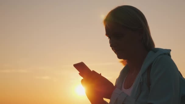 Silhuetten av en medelålders kvinna med en smartphone i solnedgången — Stockvideo