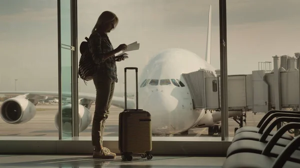 Eine Frau mit Pass und Bordkarte steht vor dem riesigen Fenster, hinter dem das Flugzeug zu sehen ist. Beginn der Reise — Stockfoto