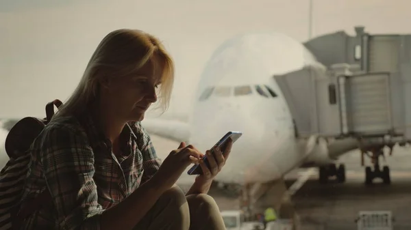 Mujer viajero utiliza un teléfono inteligente en la terminal del aeropuerto en el fondo de un gran avión de pasajeros fuera de la ventana —  Fotos de Stock