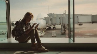 Bir kadın yolcu pencere olarak havaalanı terminal, kullanır bir smartphone oturur. Pencerenin dışında çok büyük bir uçak fonunda