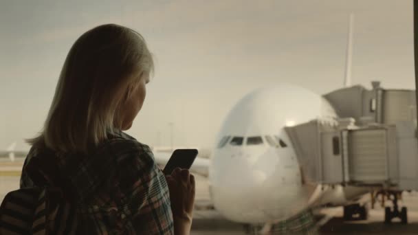 Женщина использует смартфон в терминале аэропорта на фоне большого авиалайнера за окном. Вид сзади — стоковое видео