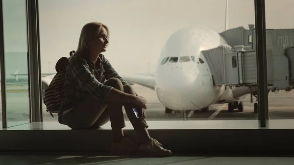 Una donna single è seduta sul davanzale della finestra nel terminal dell'aeroporto in mezzo a un enorme aereo di linea. Solitudine e concetto di separazione — Foto Stock
