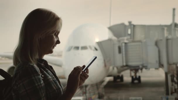 Seitenansicht der Frau benutzt ein Smartphone im Flughafenterminal auf dem Hintergrund eines großen Flugzeugs vor dem Fenster. — Stockvideo