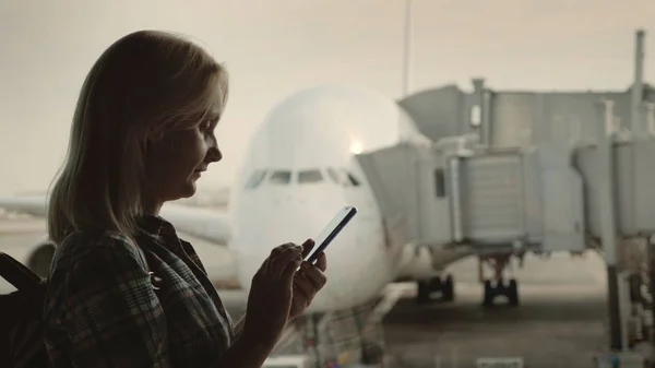 Zijaanzicht van de vrouw maakt gebruik van een smartphone in de luchthaventerminal, op de achtergrond van een groot passagiersvliegtuig buiten het raam. — Stockfoto