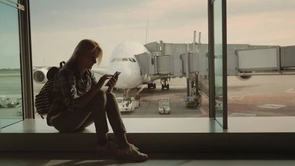 Una pasajera se sienta en el alféizar de la ventana en la terminal del aeropuerto, utiliza un teléfono inteligente. Contra el telón de fondo de un enorme avión de pasajeros fuera de la ventana —  Fotos de Stock