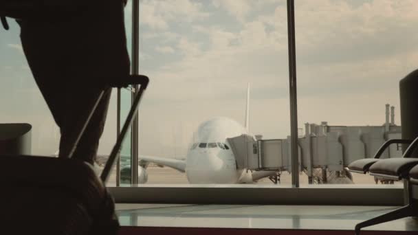 荷物を持つ女性は、空港ターミナルの大きな窓に行きます。窓の外は美しい旅客機を見ることができます。旅を見越して — ストック動画