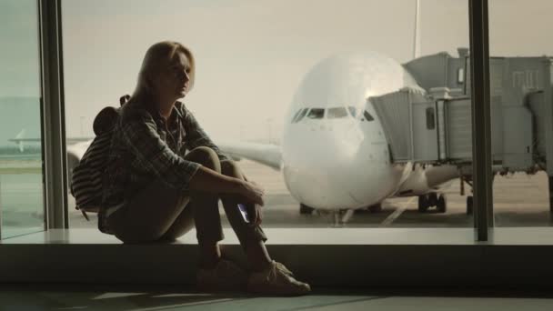 Одинокая женщина сидит на подоконнике в терминале аэропорта посреди огромного авиалайнера. Концепция одиночества и расставания — стоковое видео