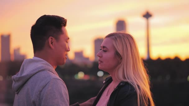 Többnemzetiségű fiatal pár beszél a hídon. A háttérben egy nagy város, és egy gyönyörű naplemente — Stock videók