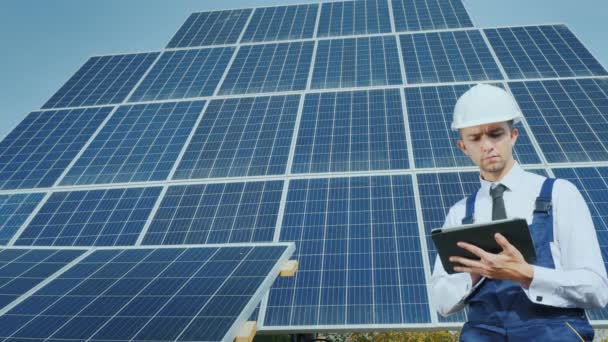 A young businessman in a white helmet uses a tablet on the background of solar panels — Stock Video