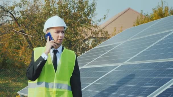 Pracovník v přilbě vejde po řadě solárních panelů, hovoří do telefonu. Steadicam shot — Stock video