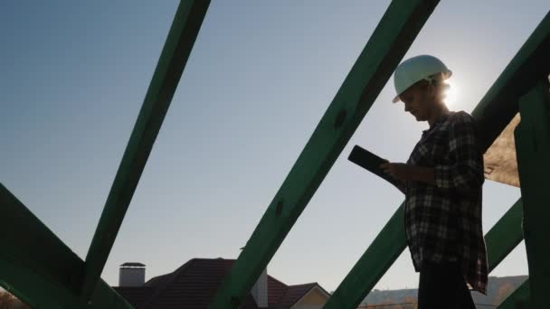 Ingenjören arbetar med en surfplatta på taket till stugan. Kontrollerar kvaliteten på byggnationen — Stockvideo