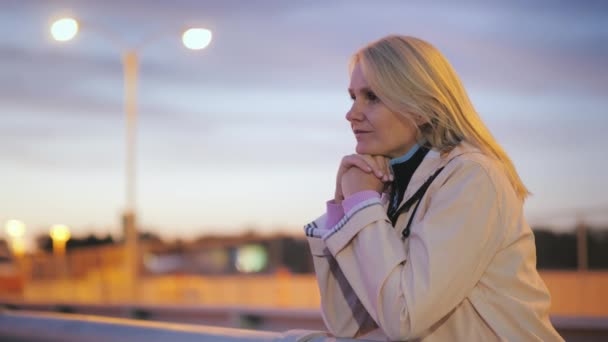 Orta yaşlı bir kadın bir köprü korkuluk, üzerinde duruyor ve düşünceli bir gece şehirde mesafe içine görünüyor — Stok video