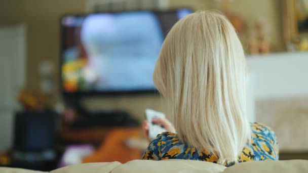 Una mujer está viendo la televisión en la sala de estar, vista trasera. Sostiene un control remoto — Vídeos de Stock