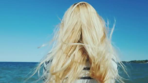 Ξανθιά γυναίκα με μακριά μαλλιά κοιτάζει τη θάλασσα. Άνεμος βολάν τα μαλλιά της — Αρχείο Βίντεο