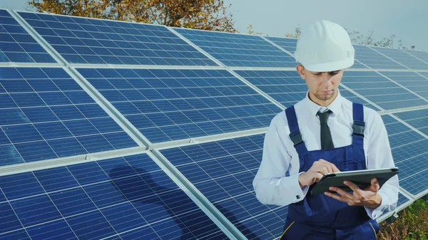 Un ingeniero con casco y mono usa una tableta en el fondo de una central solar —  Fotos de Stock