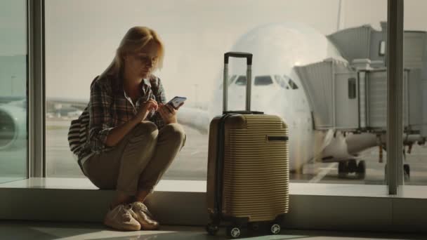 Um jovem turista com uma mala sobre rodas esperando para embarcar no avião, usando um smartphone — Vídeo de Stock