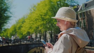 Bir kadın bir smartphone içinde elleri ve kulaklık ile bir açık bahar gününde Amsterdam'da .güzel bir kanalınkıyısında duruyor