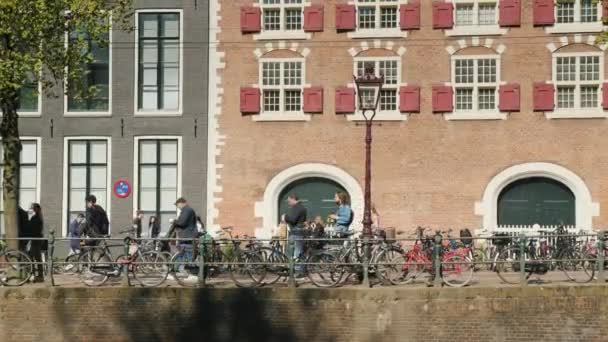Barceldam, Holanda, maio de 2018: pedestres e ciclistas estão correndo sobre seus assuntos em um dos canais de Amsterdã contra o pano de fundo de um típico edifício de tijolos para esta cidade . — Vídeo de Stock