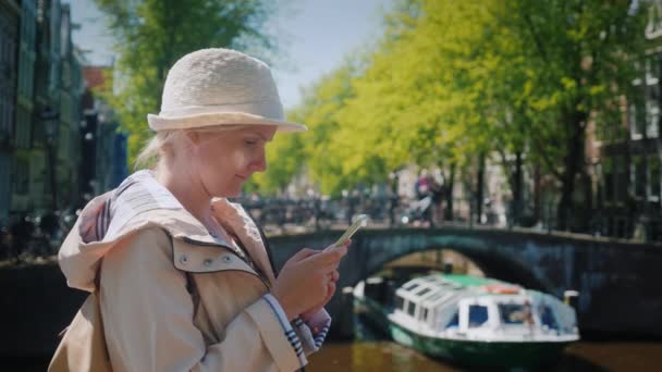 魅力的な女性は、アムステルダムの美しい運河の 1 つの近くのスマート フォンを使用します。オランダの観光 — ストック動画