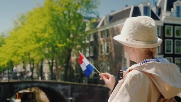 Mujer joven y elegante con la bandera de los Países Bajos se encuentra en el canal en el fondo de los edificios antiguos. Turismo en los Países Bajos — Vídeos de Stock