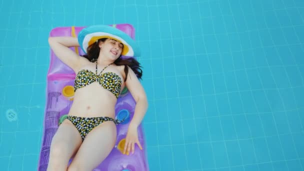 Una chica bonita de la construcción en un traje de baño y un sombrero de ala ancha brillante se relaja en un colchón inflable en una piscina con agua azul clara — Vídeo de stock