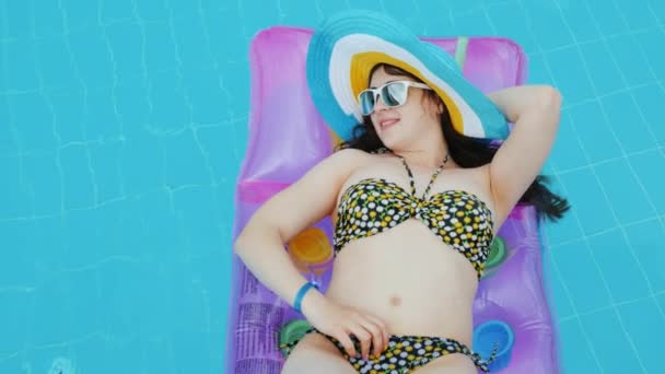 Ein junges Mädchen im Bikini und mit coolem, stylischem Hut und Sonnenbrille sonnt sich auf einer aufblasbaren Matratze in einem Pool in einem Badeort — Stockvideo