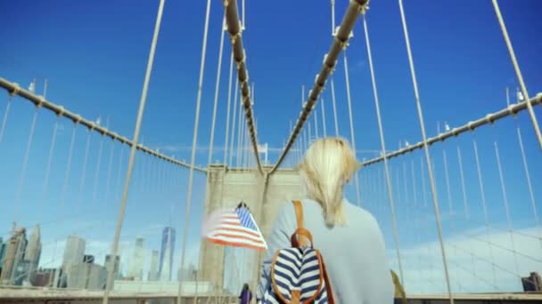 Een toerist met een vlag van Amerika en een backpacker is een wandeling langs de beroemde Brooklyn Bridge in New York. Amerika reizen concept — Stockvideo