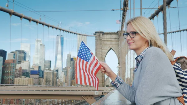Una mujer con la bandera de Estados Unidos en la mano se para en el puente de Brooklyn con vistas a Manhattan. Turismo en los Estados Unidos — Foto de Stock