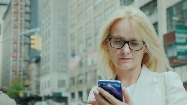 Attractive woman in glasses is looking at the screen of a smartphone. In the background are tall buildings with American flags. Manhattan, New York — Stock Video