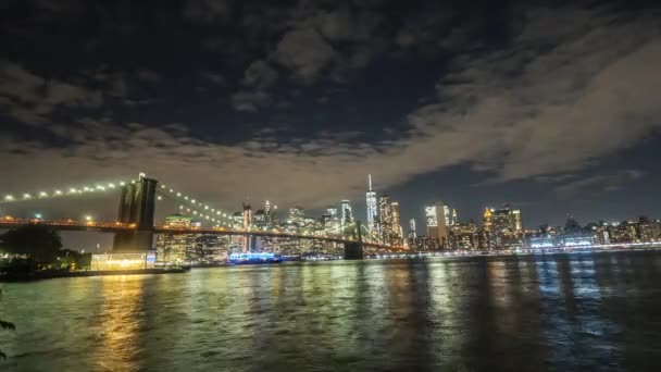 De beroemde Brooklyn Bridge's avonds schepen varen langs de rivier, op de achtergrond de lichtgevende wolkenkrabbers van Manhattan — Stockvideo