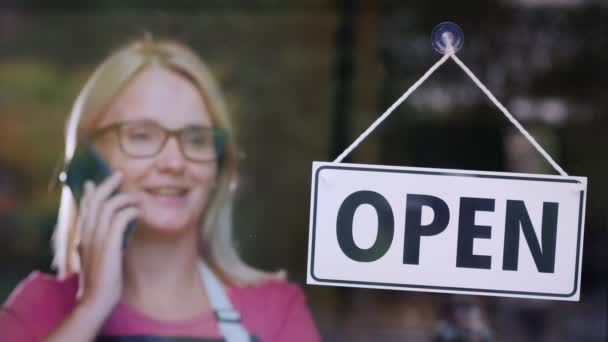 Mujer hablando por teléfono, de pie detrás de una puerta de cristal con un cartel abierto — Vídeo de stock