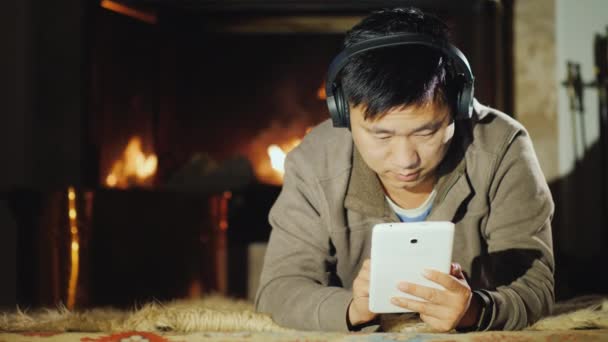Um homem asiático sério usa um tablet em casa, fica perto da lareira. Trabalho e descanso em uma casa confortável — Vídeo de Stock